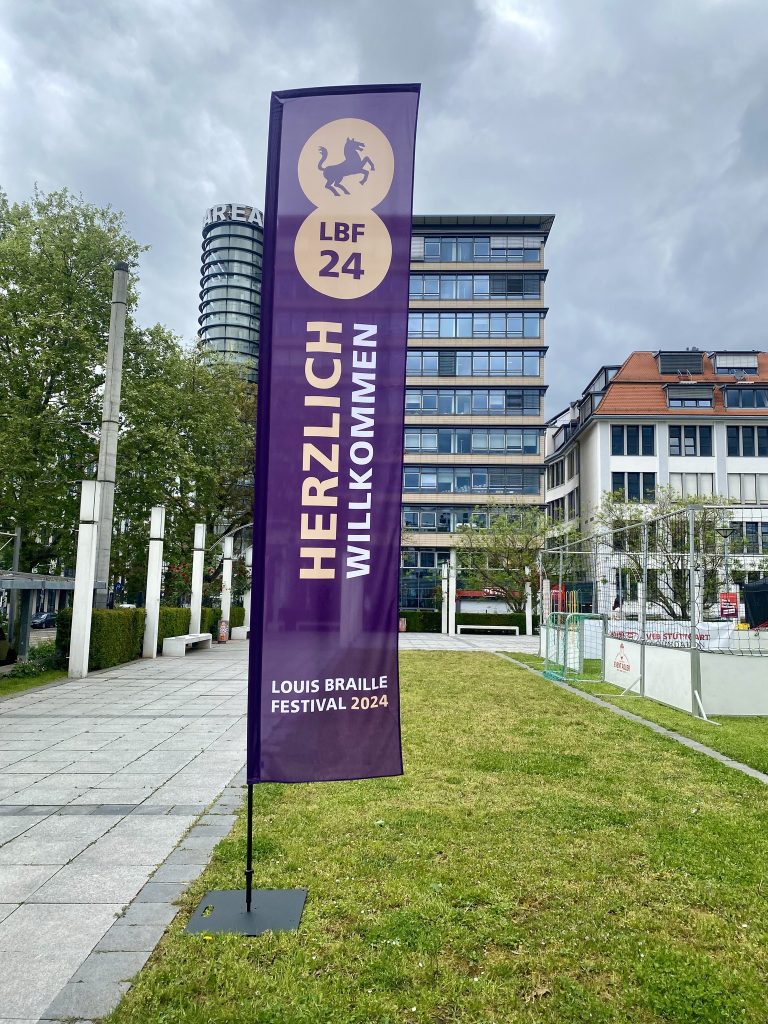 Berliner Platz/Stuttgart - Auf einer grünen Wiese steht eine Beachflag mit den Schriftzügen "Herzlich Willkommen" und "Louis Braille Festival 2024" sowie dem Logo "Pferd" und "LBF 24". Links neben der Rasenfläche ist ein breit gepflasterter Gehweg, im Hintergrund stehen große Geschäftshäuser und der Himmel ist bedeckt.