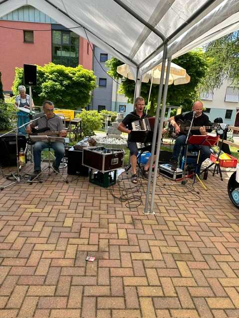 Drei Musiker spielen unter einem Pavillon, auf einem Akkordeon, einer Bass- und Akustikgitarre. Im Hintergrund sind Bäume und das Seniorenheim Rochsburg zu sehen.