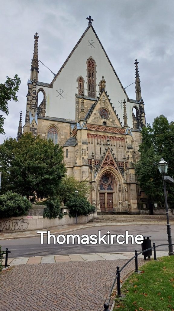 Thomaskirche Leipzig, zum Haupteingang führt eine Treppe, links und rechts stehen Bäume und Sträucher. Davor ist eine Straße, am unteren Bildrand ist ein Fuß weg mit einem Mülleimer sowie einer Laterne, und der Himmel im Hintergrund ist bewölkt.