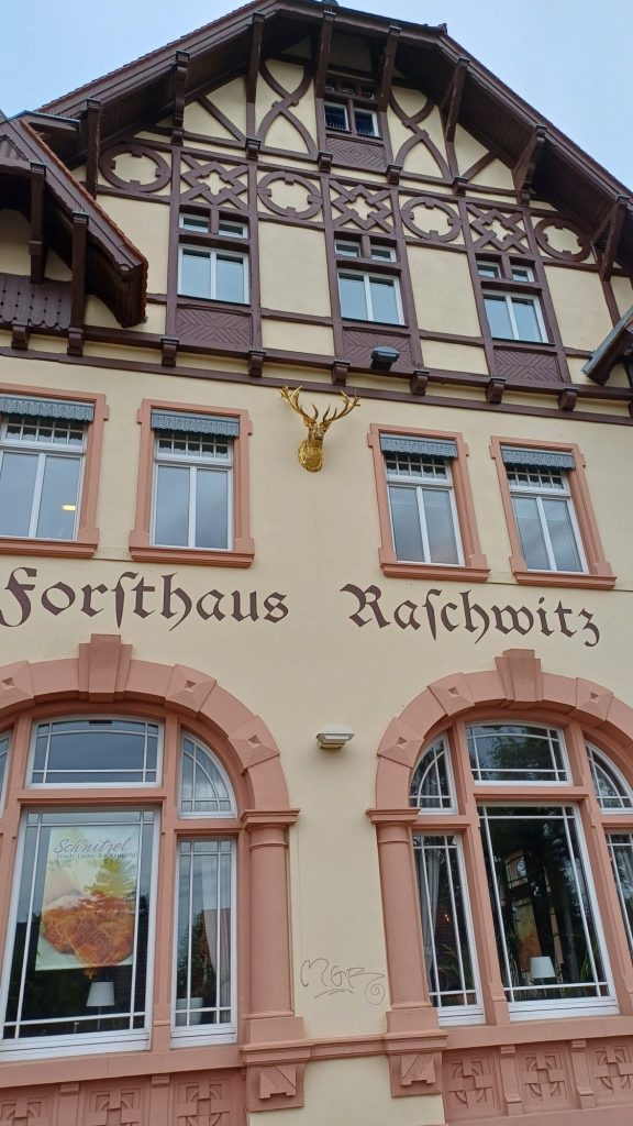 Teilabbildung des Forsthauses Raschwitz. Die ersten beiden sichtabren Etagen haben eine pastellrote Fensterlaibung, die wie obersten Etagen eine dunkelbraune Holzlaibung und es ist ein goldener Hirschkopf an der Fassade befestigt. Der Himmel im Hintergund ist hellblau.