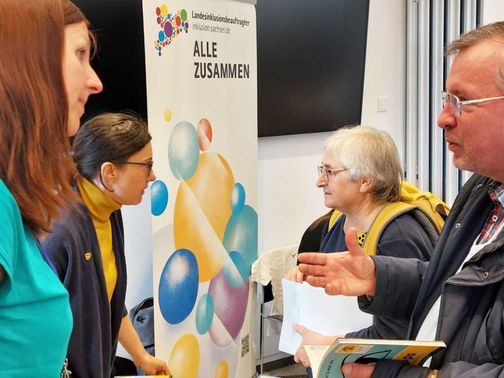 Am Stand des Landesinklusionsbeauftragten stehen zwei Besucher und zwei Vertreterinnen und unterhalten sich. Im Hintergrund steht das Roll UP von inklusion.sachsen.de, welches bunte ausgefüllte Kreise und den Schriftzug "Landesinklusionsbeauftragter, inklusion.sachsen.de, Alles zusammen" abbildet.