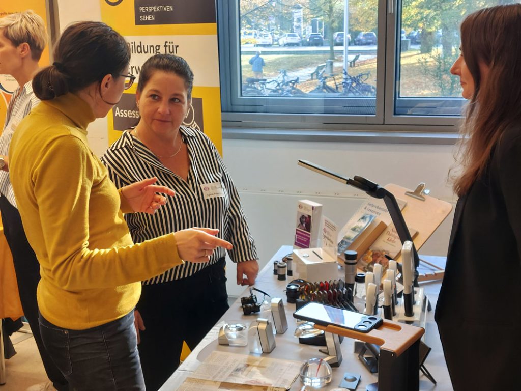 Drei Frauen unterhalten sich am Stand von Sehhilfen / Low Vision (ein Angebot des BSVS) vor ihnen, auf dem Tisch liegen verschiedene Sehhilfen.