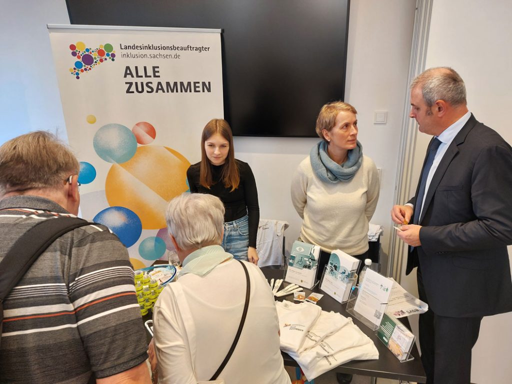 Am Stand des Inklusionsbeauftragten stehen Herr Welsch, zwei seiner Mitarbeiterinnen und zwei Besucher, welche sich unterhalten.