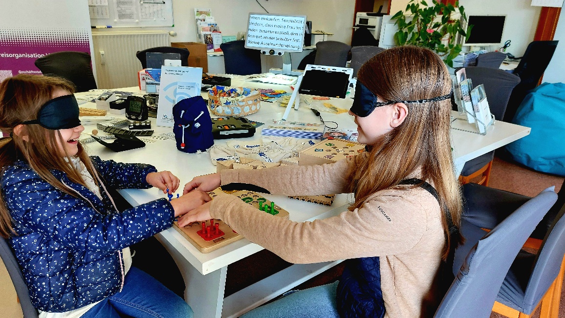 Zwei Mädchen spielen mit verbundenen Augen Mensch ärgere dich nicht. Im Hintergrund ein Tisch mit Hilfsmitteln für Blinde und Sehbehinderte.