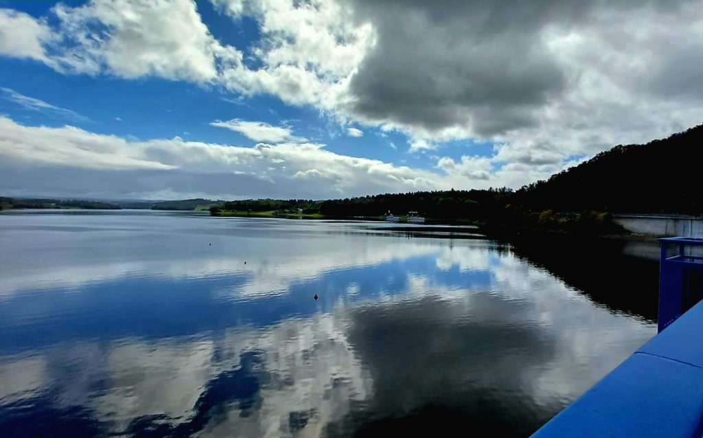 Blick von der Sperrmauer auf die randvolle Talsperre. Der Himmel spiegelt sich im Wasser.