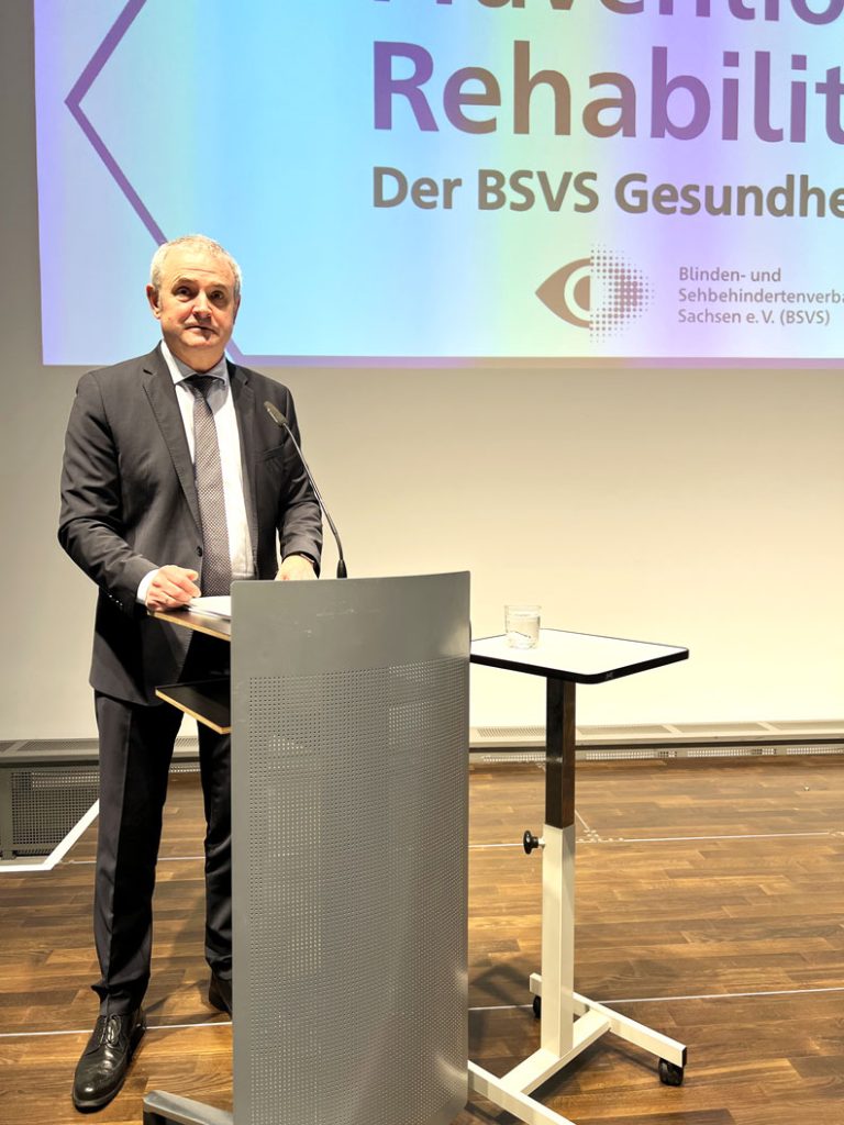 Herr Welsch steht auf der Bühne am Pult und begrüßt die Gäste zum Gesundheitstag des BSVS. Im Hintergrund an der Wand ist ein Teil des Standbildes zum Gesundheitstag zu sehen.