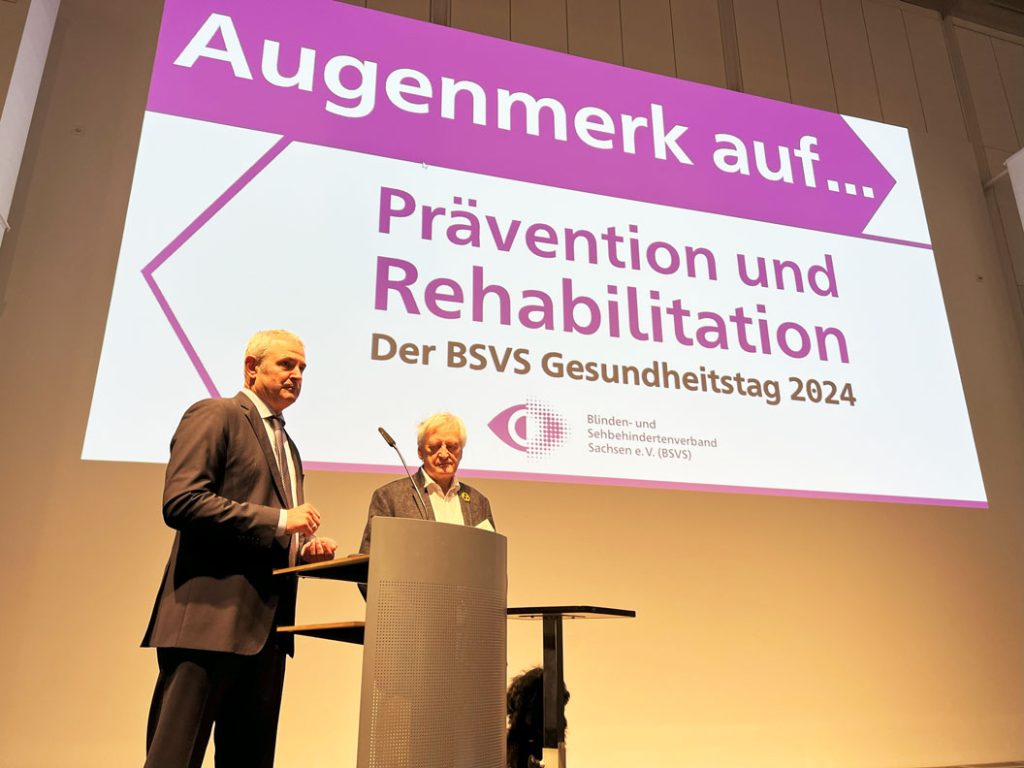 Herr Welsch und Herr Schneider stehen gemeinsam auf der Bühne und halten die Abschlussrede zum Gesundheitstag. Im Hintergrund ist das Standbild zum Gesundheitstag an die Wand projiziert.