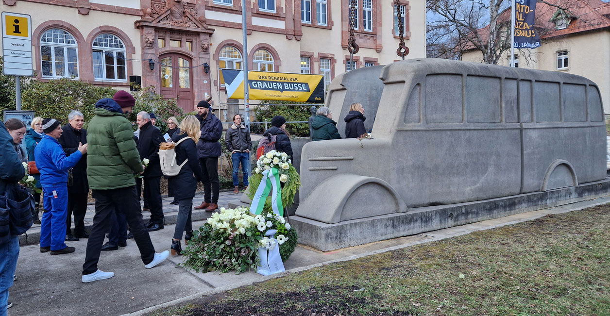 Auf der rechten Seite ist das Denkmal, davor liegen Kränze und es stehen Menschen am „Grauen Bus“. Im Hintergrund ist ein SFZ Gebäude.